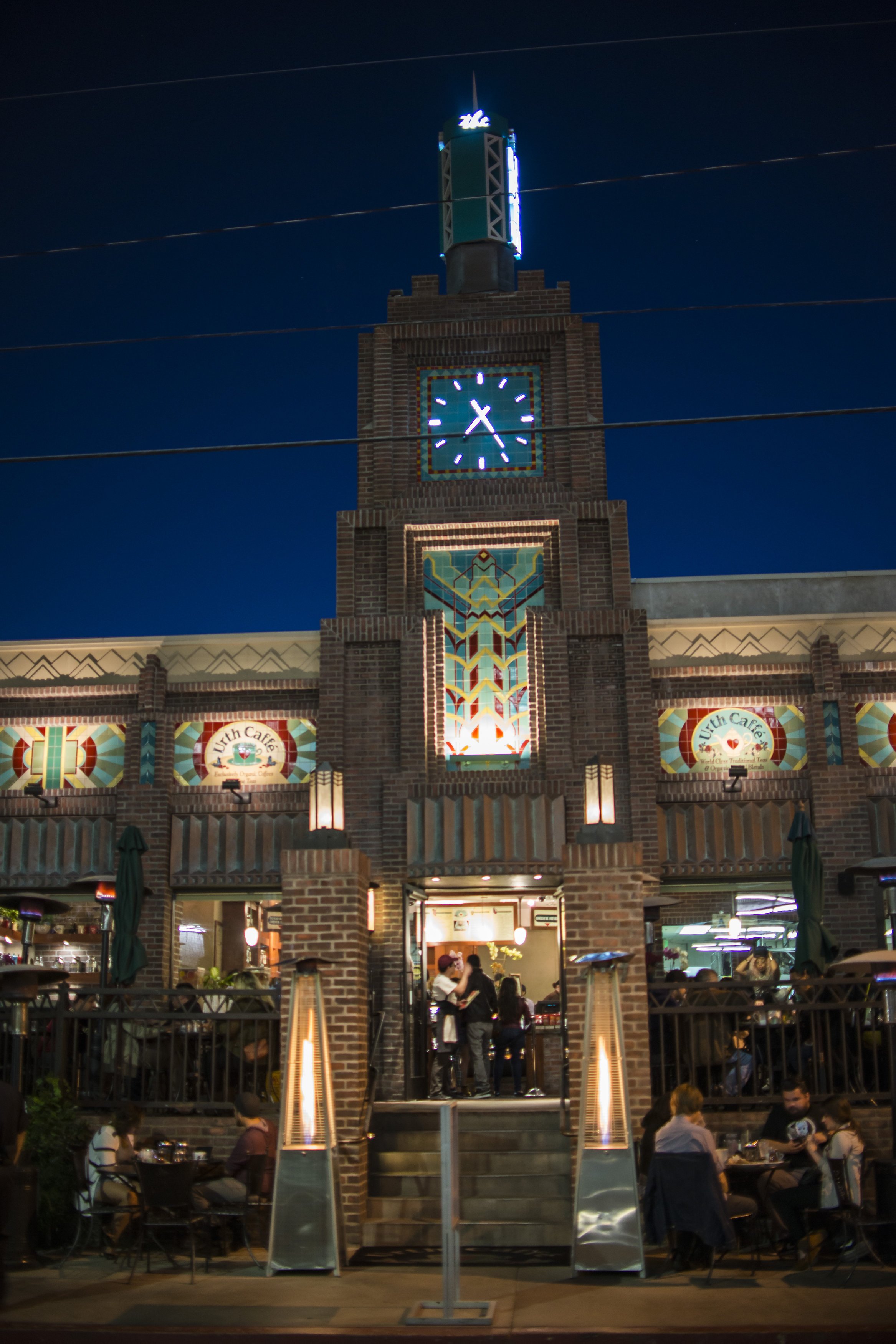 A nighttime view of a hotel's exterior, showing a clock tower, brick facade, and outdoor seating area with people dining.