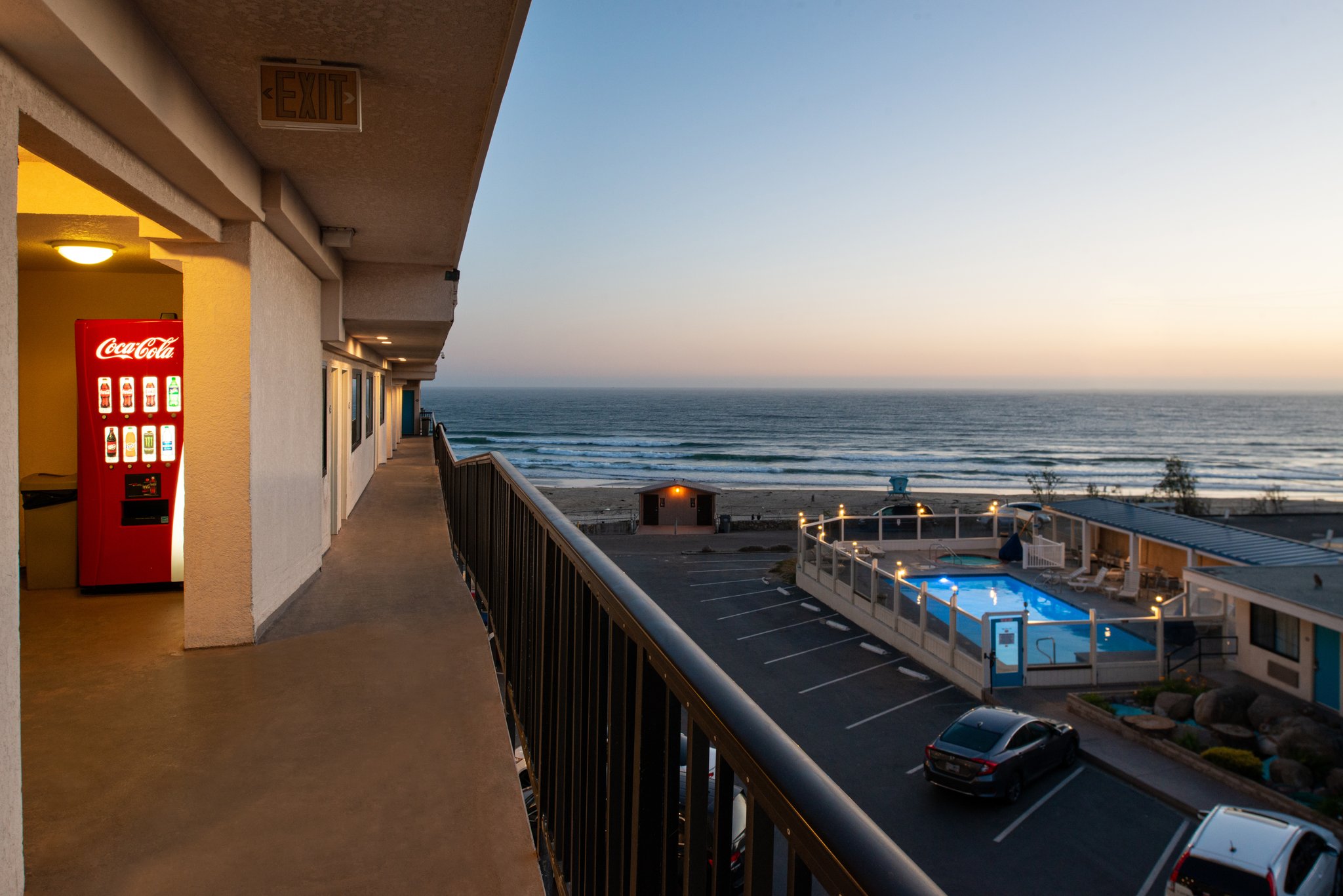 A balcony overlooking the ocean, pool, and parking lot of a hotel.