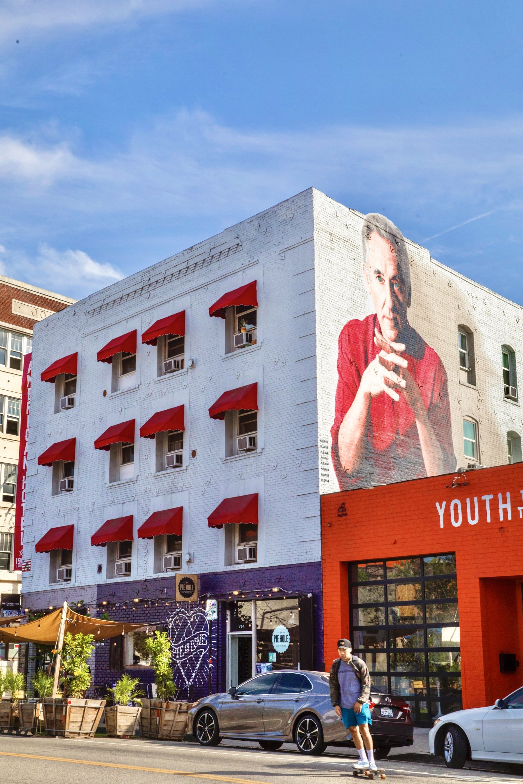 A white brick building with a large mural of a man on the side, red awnings on the windows, and a blue sky in the background.