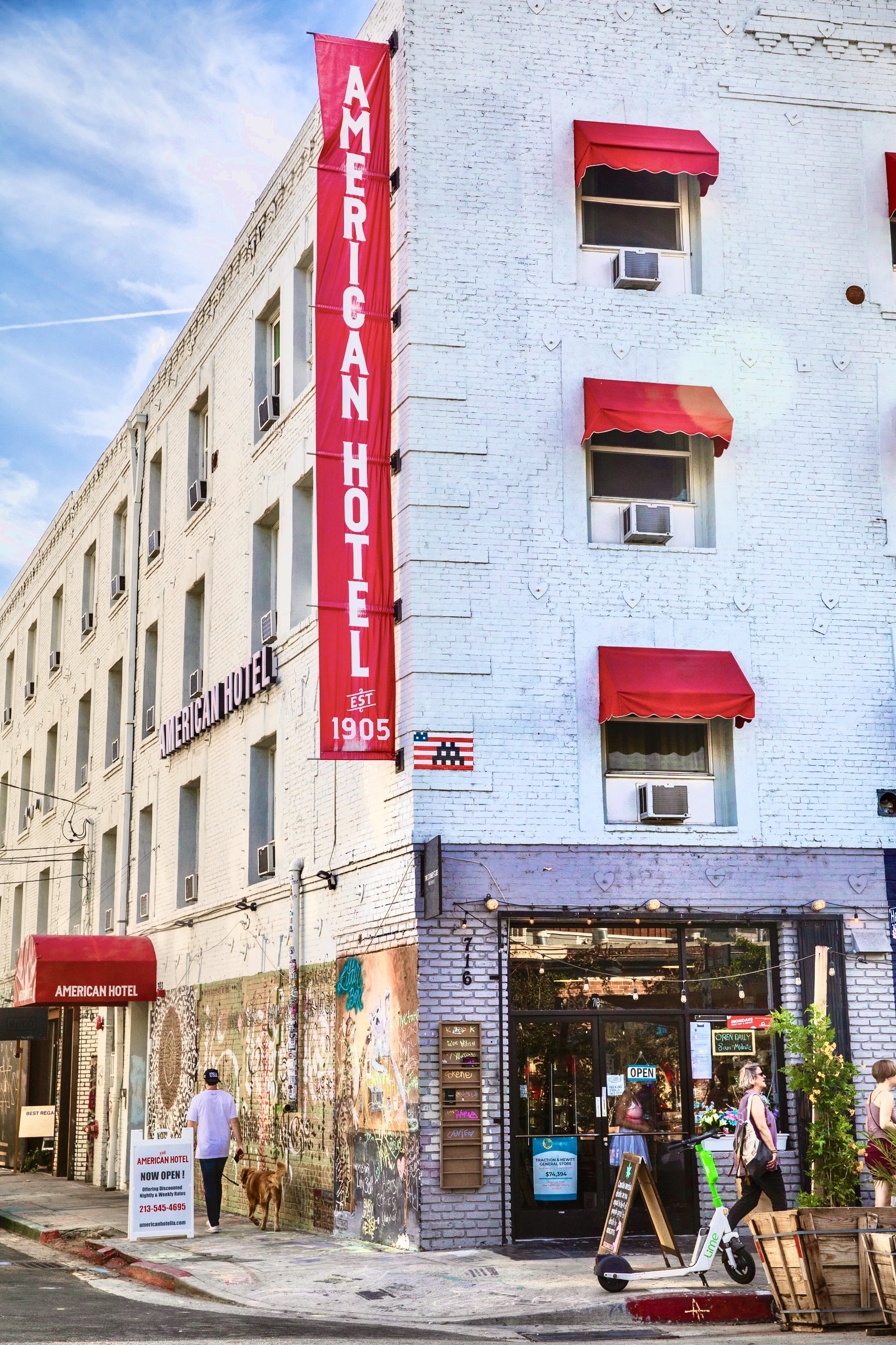 The exterior of a white brick hotel with a red banner and awnings, featuring a storefront and sidewalk activity.